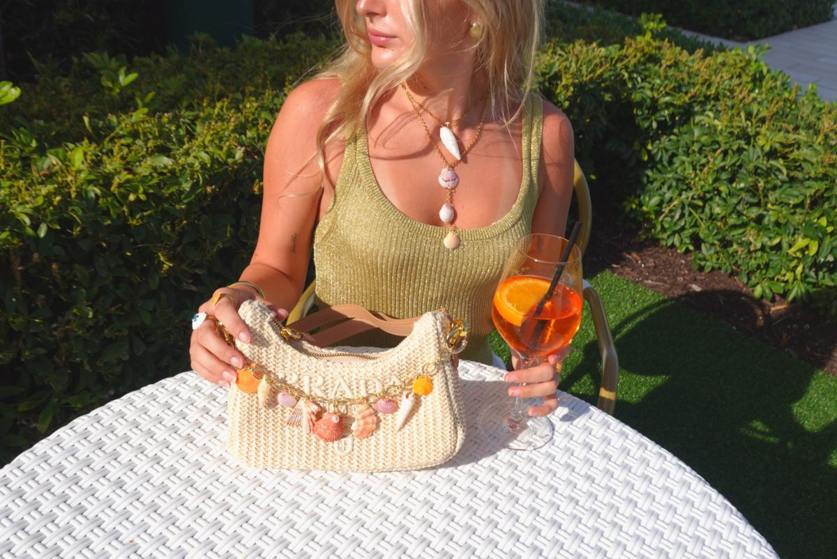 Shell charm chain styled on a straw Prada bag beside an Aperol spritz