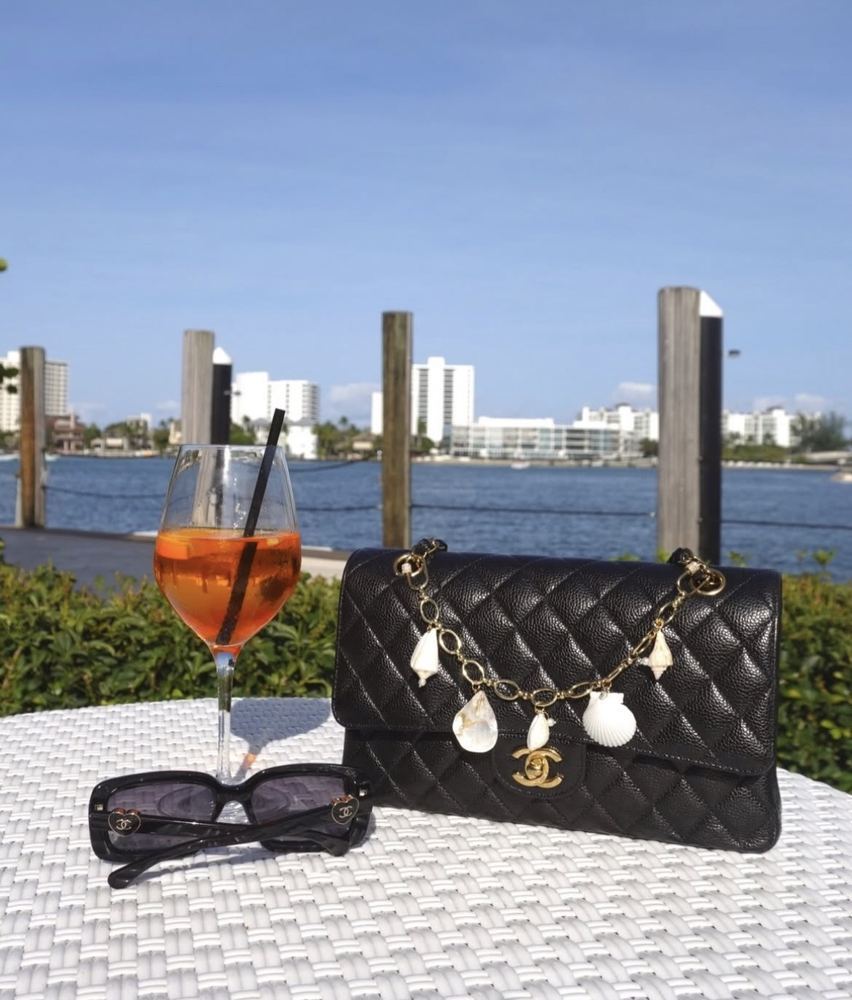 Shell charm on a black quilted bag beside an Aperol spritz and sunglasses on the water